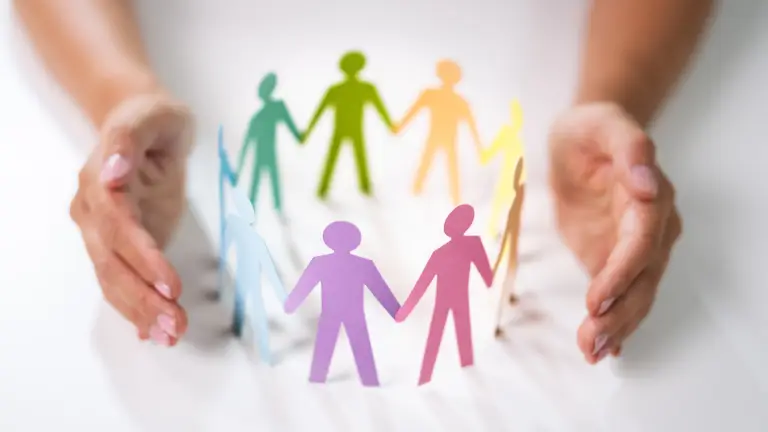 Paper cut-outs of people holding hands in a circle, surrounded by real human hands — symbolising unity, wellbeing, and inclusion.