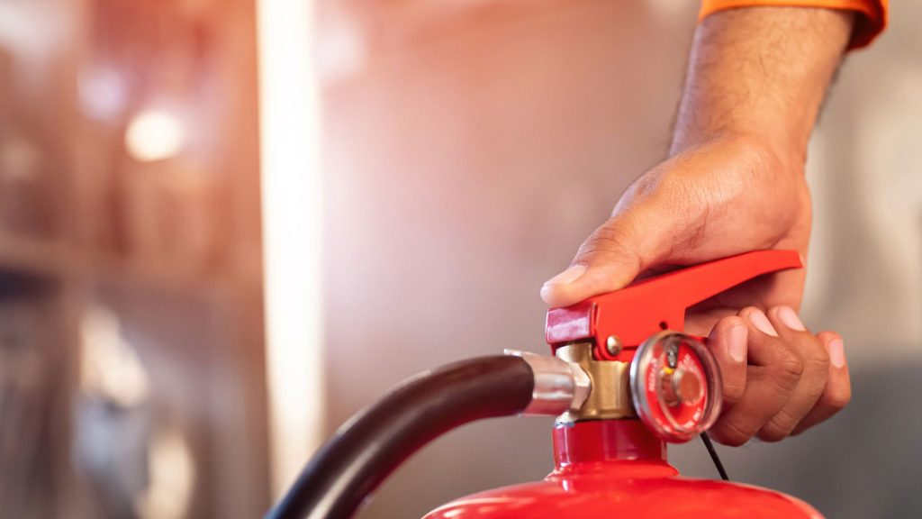 A close-up photograph of a red fire extinguisher mounted on a wall, emphasising its accessibility and readiness. The image highlights the importance of fire safety measures, linking to a discussion on Fire Risk Assessments (FRA) and ensuring compliance in workplaces and public spaces.