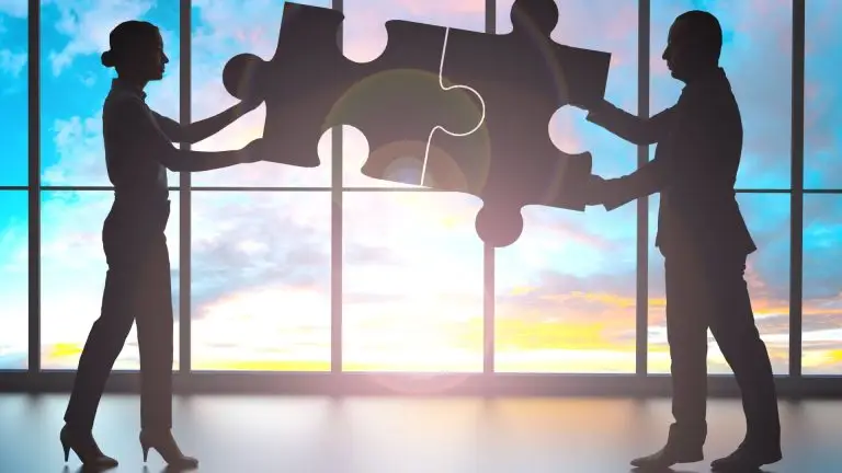 Silhouettes of a man and woman holding two puzzle pieces joined together against a bright window — representing collaboration and trusted partnerships.