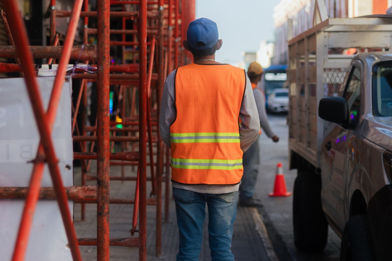 Construction worker in a neon orange safety vest and blue cap stands on a city street, scaffolding to the left and a truck to the right, highlighting workplace safety leadership in action.