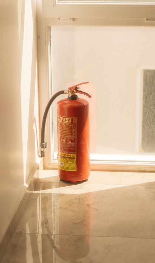 Red fire extinguisher placed indoors beside a window, ready for use, reflecting on polished floor.