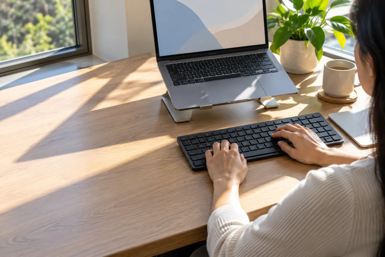 A person types on a keyboard at a desk with a laptop on a stand.